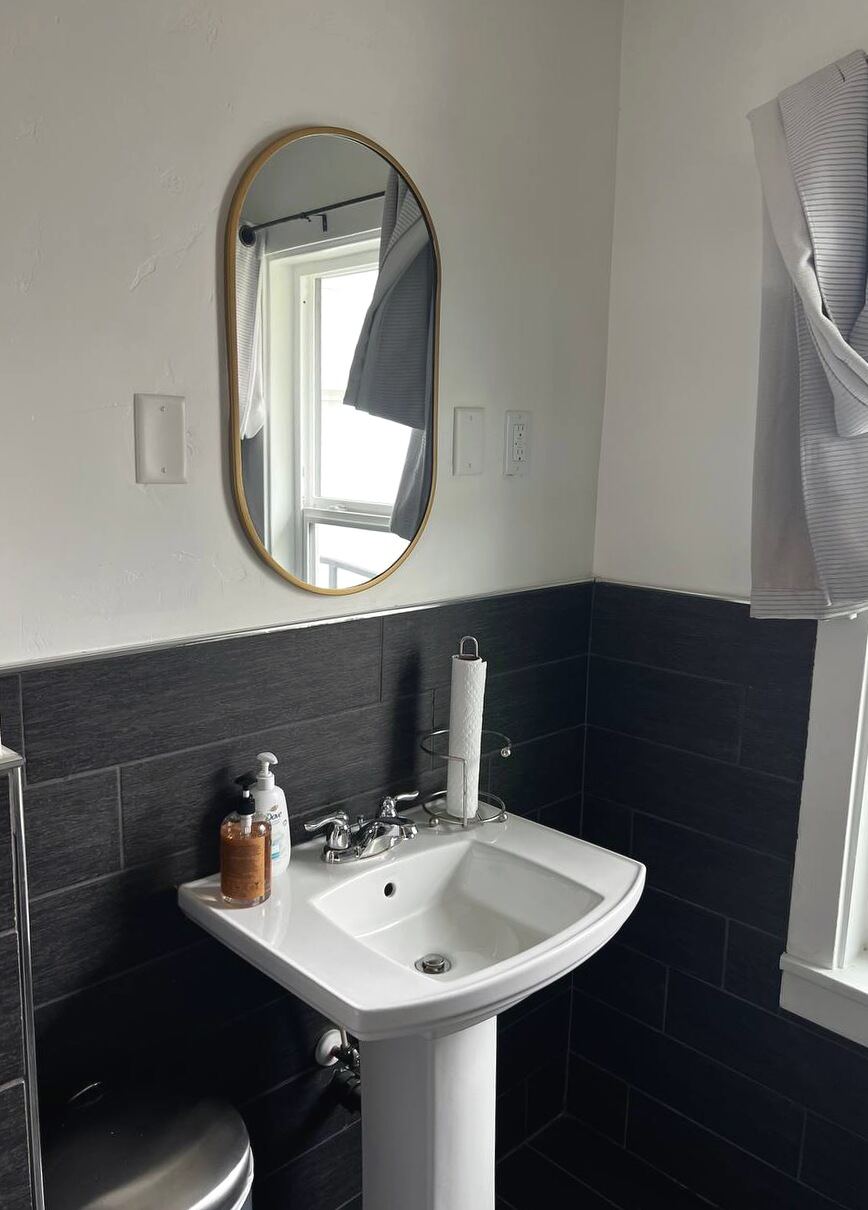 Bathroom vanity with pedestal sink, brass-framed oval mirror and full-height charcoal tile